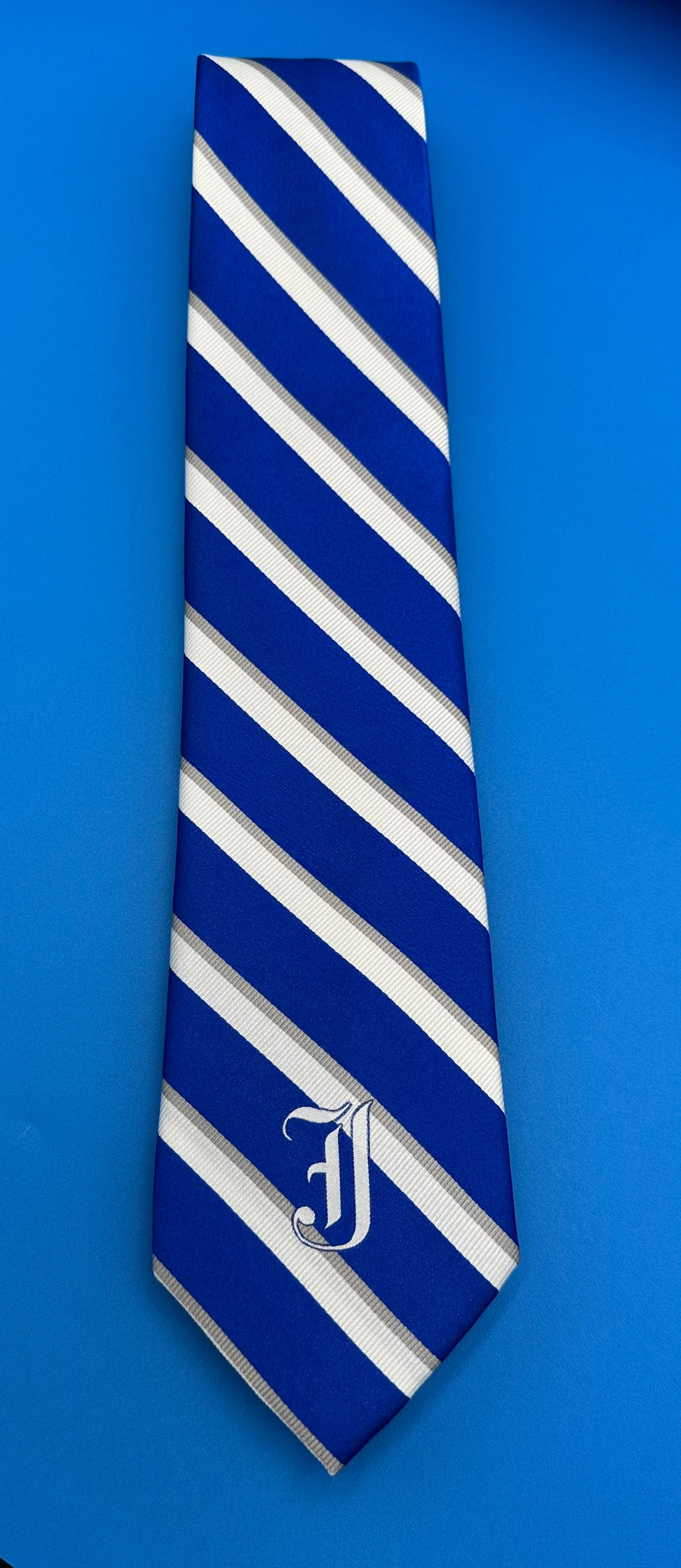 NOLA Couture.  Our newest Jesuit tie is 100% woven silk. A traditional royal blue/white/grey diagonal stripe with the embroidered English J logo.  58" length. Dry Clean Only.